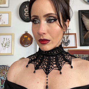 May include: A black crocheted choker necklace featuring an intricate web-like pattern, a high collar design, and dangling black teardrop beads. The necklace is accented with small black beads and a central silver-toned filigree bead cap.