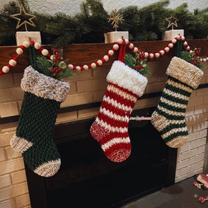 Peut inclure: Trois chaussettes de Noël crochetées suspendues en rangée. La chaussette de gauche est vert foncé avec un talon et une pointe beiges et un revers beige texturé. Celle du milieu a des rayures rouges et blanches avec un revers blanc duveteux. Celle de droite a des rayures vertes et blanches avec un revers beige duveteux. Chacune contient des branches de pin vertes et des baies rouges.