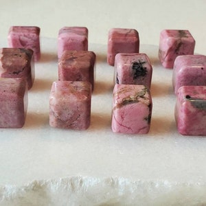 May include: A collection of 14 pink and white polished Rhodonite cubes. The cubes are arranged in a grid pattern on a white surface.