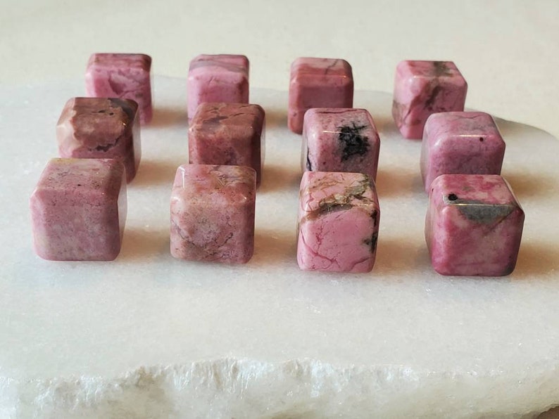 May include: A collection of 14 pink and white polished Rhodonite cubes. The cubes are arranged in a grid pattern on a white surface.