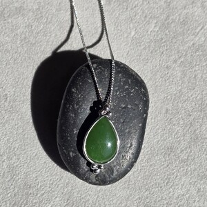 May include: A silver necklace with a teardrop-shaped, green jade pendant. The pendant is set in a silver bezel and hangs from a delicate chain. The necklace is displayed on a dark gray stone against a light gray background.