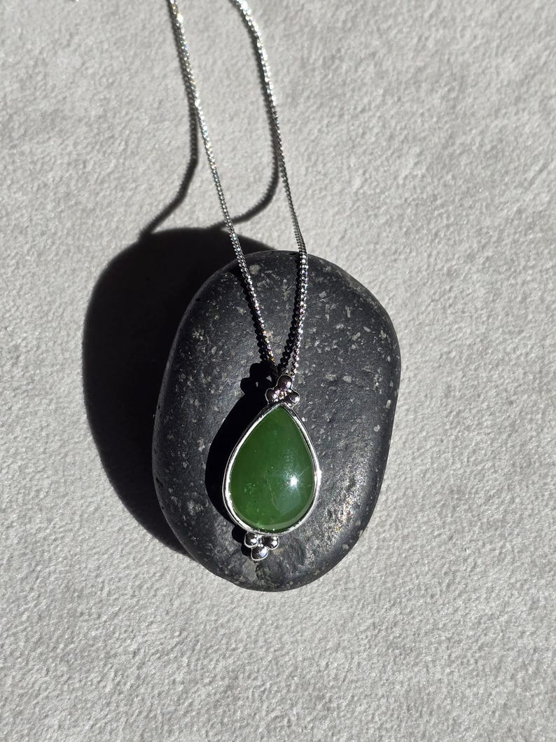 May include: A silver necklace with a teardrop-shaped, green jade pendant. The pendant is set in a silver bezel and hangs from a delicate chain. The necklace is displayed on a dark gray stone against a light gray background.