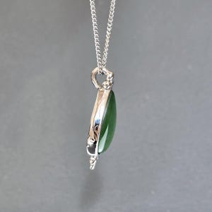 May include: A silver pendant necklace featuring a teardrop-shaped, polished green stone. The stone is encased in a silver setting and hangs from a delicate silver chain. The pendant has a detailed design at the top and bottom.