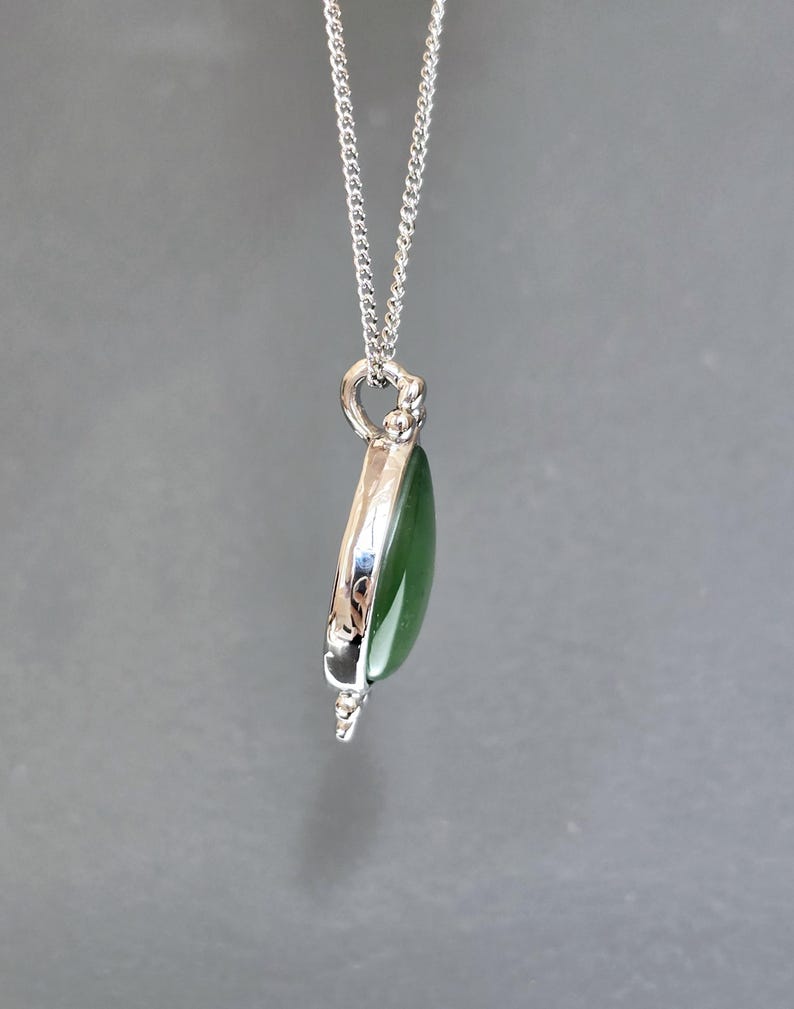 May include: A silver pendant necklace featuring a teardrop-shaped, polished green stone. The stone is encased in a silver setting and hangs from a delicate silver chain. The pendant has a detailed design at the top and bottom.