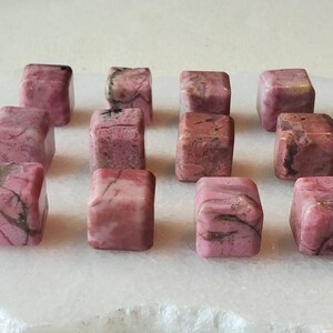 May include: A collection of 12 pink and white tumbled rhodochrosite stones, each cut into a cube shape. The stones are arranged in rows on a white surface.
