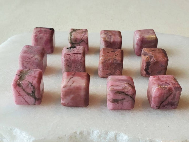 May include: A collection of 12 pink and white tumbled rhodochrosite stones, each cut into a cube shape. The stones are arranged in rows on a white surface.