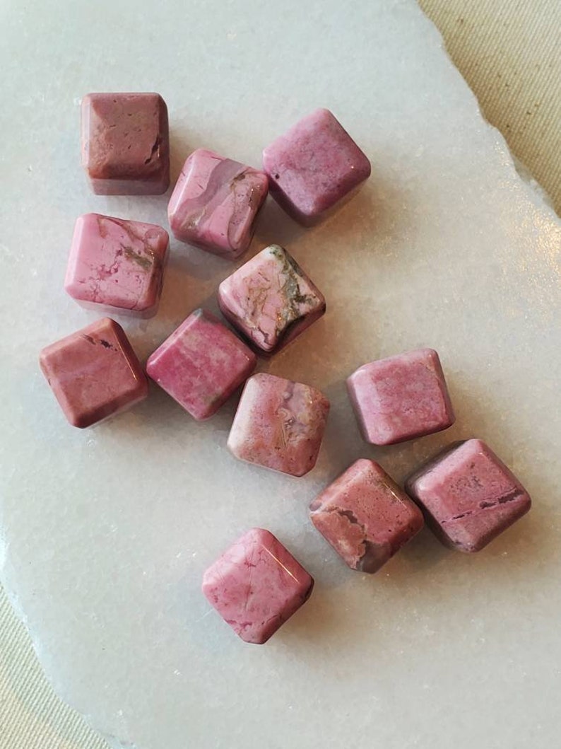 May include: A collection of pink and white polished rhodochrosite gemstone cubes. The cubes are various sizes and have a natural, rough texture.