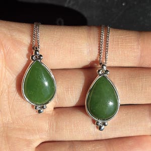 May include: Two jade teardrop pendants on silver chains. The green jade stones are set in a silver bezel with decorative silver accents. The pendants are displayed on a light-toned background.