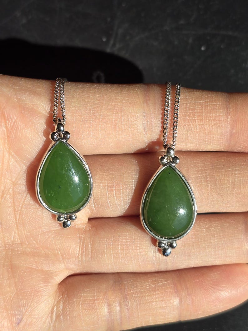 May include: Two jade teardrop pendants on silver chains. The green jade stones are set in a silver bezel with decorative silver accents. The pendants are displayed on a light-toned background.