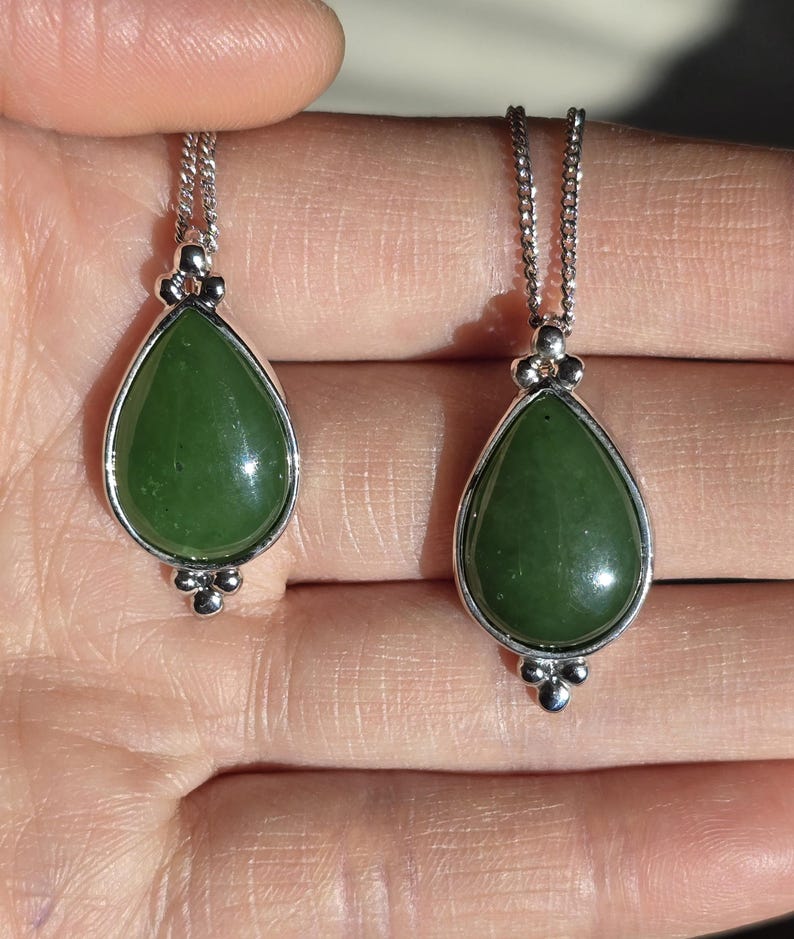May include: Two jade teardrop pendants on silver chains. The green jade stones are set in silver bezels with decorative silver accents. The pendants are displayed against a neutral background.