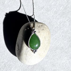 May include: A silver necklace with a teardrop-shaped green jade pendant. The pendant is set in a silver bezel and hangs from a delicate chain. The necklace is displayed on a light-colored stone.