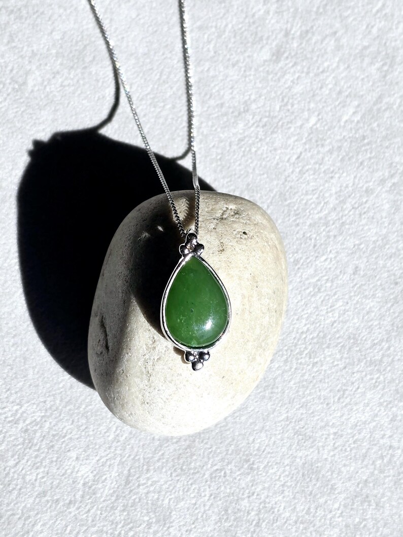 May include: A silver necklace with a teardrop-shaped green jade pendant. The pendant is set in a silver bezel and hangs from a delicate chain. The necklace is displayed on a light-colored stone.