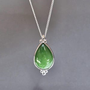 May include: A silver necklace featuring a teardrop-shaped pendant with a vibrant green stone. The stone is encased in a silver setting and hangs from a delicate chain. The pendant has a decorative design at the top and bottom.