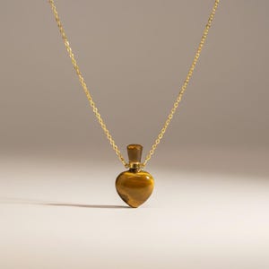 May include: A gold-colored necklace with a heart-shaped pendant. The pendant is made of a brown, tiger's eye stone. The necklace chain is delicate and hangs from the pendant's top. The pendant has a small, brown stopper.