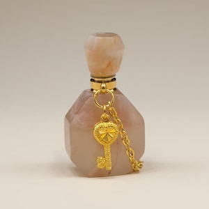May include: A faceted, peach-colored perfume bottle with a gold-colored key charm hanging from a chain. The bottle has a matching peach-colored stopper. The key charm is heart-shaped.