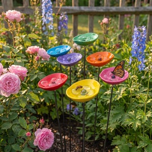 Puede incluir: Un conjunto de coloridos bebederos para pájaros de cerámica sobre tallos metálicos, dispuestos en un jardín. Los bebederos son de varios colores, incluyendo rojo, amarillo, rosa, naranja, morado, azul y verde. Una mariposa y una abeja son visibles en dos de los bebederos.
