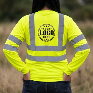 Puede incluir: Una camiseta de manga larga de color amarillo neón de alta visibilidad con bandas reflectantes plateadas horizontales y verticales. La espalda muestra un gráfico circular negro con el texto YOUR LOGO HERE y estrellas entre las bandas.