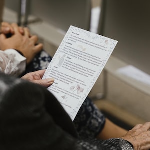 May include: A person holds a printed wedding program with text and decorative floral borders. The program is white with black text and illustrations. The person is wearing a patterned top and a ring.