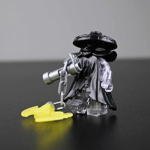 May include: A black minifigure with a wide-brimmed hat and a tattered cloak. The figure has a silver chain and a yellow accessory. The figure's legs are transparent. The background is a solid gray.