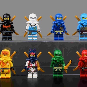 May include: Eight colorful Lego Ninjago minifigures are displayed on a clear acrylic shelf. Each figure is a different color, including black, white, blue, and red, and has a matching head covering and gold-colored weapons.