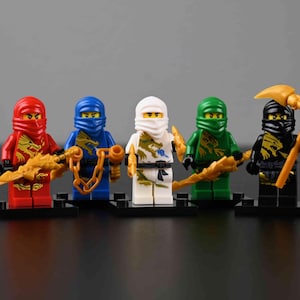 May include: Five Lego Ninjago minifigures in red, blue, white, green, and black, each with a matching head covering. They hold golden weapons, including a sword, chain, and scythe. The figures stand on black bases.