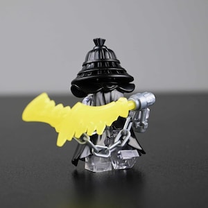 May include: A black minifigure with a black hat and cloak, holding a yellow weapon. The figure has a silver chain and a silver gun-like accessory. The figure is on a clear base.