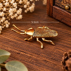 May include: A gold-toned metal beetle figurine with textured wing covers and two long antennae. The insect-shaped object features a detailed, segmented body and legs, with a measurement indicator clearly showing a length of 5.5 cm.