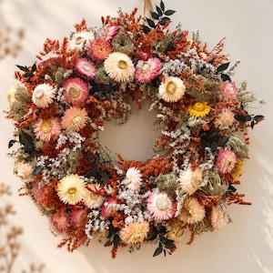 May include: A circular wreath composed of dried flowers and foliage in shades of rust orange, cream, pale pink, and dark green. The arrangement features numerous strawflowers, fine white filler plants, and dark green leaves on a base.