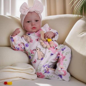 May include: A baby wearing a white onesie with a bunny and floral print, a matching headband with a bow, and holding a wooden toy. The onesie has pink and purple flowers and butterflies. The baby is sitting on a cream-colored couch.