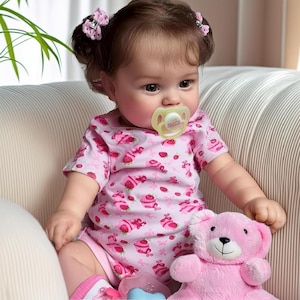 May include: A baby doll with brown hair and eyes, wearing a pink onesie with a strawberry and cupcake pattern. The doll has a pacifier and is holding a pink teddy bear. The doll is sitting on a beige couch.