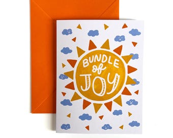 Op de afbeelding: Een witte wenskaart met een centrale gele en oranje zonne-illustratie met de tekst BUNDLE of JOY in het wit. De kaart is omringd door kleine blauwe wolkjes en oranje driehoekjes, vergezeld door een effen oranje envelop.