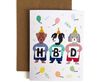 Op de afbeelding: Een witte verjaardagskaart met drie geïllustreerde honden in kleurrijke feesthoedjes en truien, die elk een bordje vasthouden met de letters H, B en D. De kaart is versierd met gekleurde ballonnen en confetti en wordt geleverd met een bruine envelop.