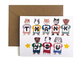 Op de afbeelding: Een witte wenskaart met een kleurrijke illustratie van acht getekende honden in twee rijen. Elke hond houdt een wit bordje met een zwarte letter vast, die samen de woorden THANK YOU vormen. Twee gele sterren staan aan de onderkant.