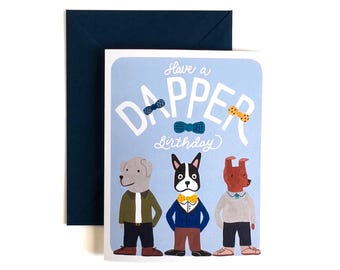 Op de afbeelding: Een lichtblauwe verjaardagskaart met drie geïllustreerde honden in truien en met strikjes. De tekst luidt Have a DAPPER Birthday. De kaart wordt geleverd met een donkerblauwe envelop. De illustratiestijl is plat en kleurrijk.