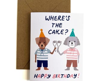 Op de afbeelding: Een witte verjaardagskaart met twee geïllustreerde honden die feesthoedjes en gestreepte shirts dragen en vorken vasthouden. De tekst luidt WHERE'S THE CAKE? en HAPPY BIRTHDAY! Een bruine envelop bevindt zich achter de kaart.