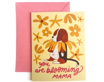Op de afbeelding: Een rechthoekige wenskaart met een lichtgele achtergrond met een bruin-witte hond die een gele bloemenkraag draagt, omringd door gele bloemillustraties en de rode tekst "you are blooming MAMA" met een roze envelop.