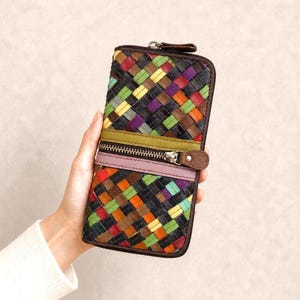 Puede incluir: Una cartera de cuero tejido multicolor con cierre de cremallera. La cartera presenta una variedad de colores que incluyen rojo, verde, naranja, morado y amarillo. La cartera está sujeta en la mano de una persona.