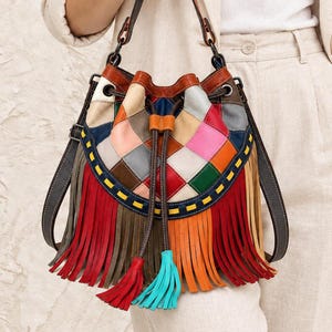 Puede incluir: Un bolso tipo saco de cuero multicolor con un diseño de parches de paneles cuadrados en rojo, naranja, verde, azul y beige. El bolso incluye un cierre de cordón con dos borlas, una correa de hombro marrón oscuro y flecos largos.