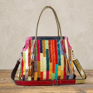 May include: A multi-coloured handbag featuring a vertical patchwork design with various textured patterns. The bag includes two braided top handles, two decorative leather tassels, and a detachable red shoulder strap with a metal buckle.