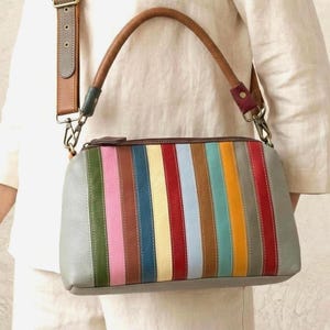 Peut inclure: Un sac à main en cuir gris avec un motif de rayures verticales colorées. Les rayures comprennent du vert, du rose, du marron, du bleu, du jaune, du rouge et de l'orange. Le sac a une poignée en cuir marron et une bandoulière amovible.