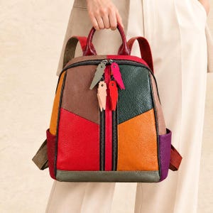 Könnte beinhalten: Ein mehrfarbiger Lederrucksack mit einem Color-Block-Design in Rot, Orange, Braun, Dunkelgrün, Lila und Grau. Die Tasche verfügt über zwei vertikale Reißverschlusstaschen auf der Vorderseite mit vier dekorativen blitzförmigen Anhängern.
