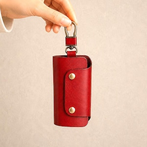 May include: A deep red leather key case featuring a smooth finish, a wrap-around flap secured by two brass-toned metal snap buttons, and a matching red leather loop attached to a swivel lobster clasp made of antique brass-toned metal hardware.