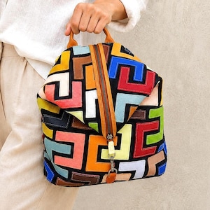Op de afbeelding: Een rugzak met een zwarte basis en een geometrisch patroon van in elkaar grijpende L-vormige patches in levendige kleuren zoals rood, blauw, geel, oranje, groen en lichtroze. De tas heeft een centrale verticale rits en een bruine leren handgreep.