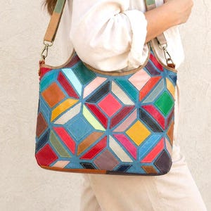 Op de afbeelding: Een kleurrijke patchwork leren tote bag met een geometrisch ontwerp. De tas heeft een verscheidenheid aan kleuren, waaronder blauw, rood, groen, geel en bruin. Hij heeft een schouderriem en een handvat aan de bovenkant. De tas is ongeveer 30 cm breed en 25 cm hoog.