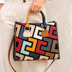 May include: A square tote bag featuring a geometric patchwork pattern of multi-colored leather pieces in red, blue, yellow, tan, and black. The bag has two distinct handles, one yellow and one dark blue, attached with gold-toned metal rings.