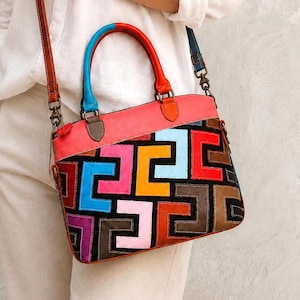 May include: A multi-colored leather handbag featuring a patchwork design of geometric shapes in shades of red, blue, yellow, pink, purple, and brown against a black background. It has a coral top panel, a bright blue handle, and a brown shoulder strap.