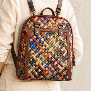 May include: A backpack featuring a multi-colored woven pattern with a grid of small squares in shades of blue, red, yellow, green, and orange. The bag includes brown leather trim, a top handle, two adjustable shoulder straps, and metal hardware.