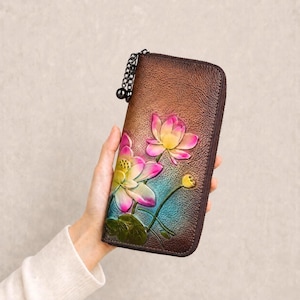 Puede incluir: Una cartera de cuero marrón con un diseño floral. La cartera presenta flores de loto rosas y amarillas en relieve con hojas verdes y detalles azules. Una cadena y una bola negras están unidas a la cremallera.