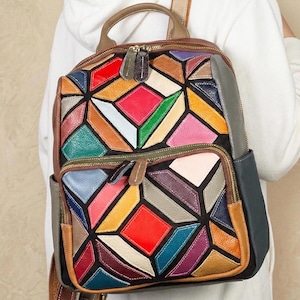 Op de afbeelding: Een kleurrijke geometrische patchwork rugzak met een handvat aan de bovenkant en twee compartimenten met ritssluiting. De tas heeft een levendige reeks kleuren, waaronder rood, geel, groen, blauw en paars. De tas heeft een bruin lederen handvat en riemen.