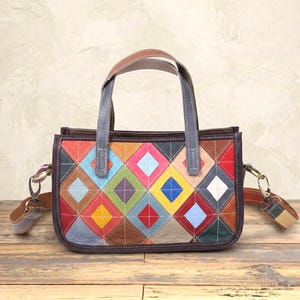 May include: A rectangular handbag featuring a multi-colored diamond patchwork pattern in shades of red, blue, yellow, green, and brown. The bag has two grey leather top handles and detachable side straps with metal lobster claw clasps.